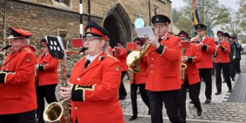 Screenshot 2023-06-13 124352 A marching band in bright red uniforms plays instruments whilst walking down a street.