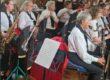 A band of musicians in white shirts plays woodwind instruments indoors with sheet music stands.